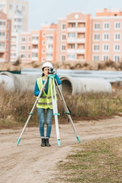 Licensed surveyor using tripod equipment to measure ground and building elevations for a flood elevation survey near residential buildings
